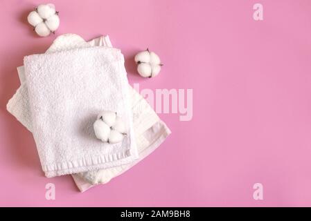 Stack of white cotton clothes and cotton flowers on pink background, flat lay, copy space. Organic cotton towels and clothes, eco lifestyle concept. Stock Photo