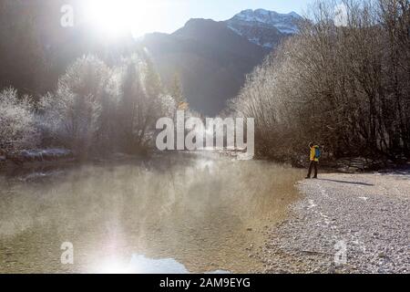 Woman in yellow jacket wearing a backpack standing by the river on the shore of lake Bohinj on a sunny misty winter day, slovenia Stock Photo
