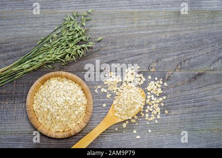 Oatmeal flakes in a bamboo bowl with a spoonful of scattered flakes and green ears of oats on an old faded gray wooden table background. The concept o Stock Photo