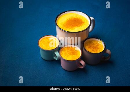 Turmeric drink in one big cup and three little cups on phantom blue background. Cups of yellow milk with turmeric powder. Detox drink. Warm beverage. Stock Photo