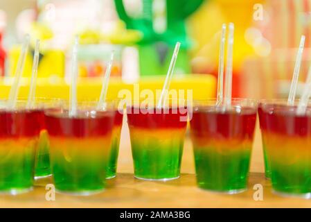 Glass with colorful striped jelly above wooden table. Rainbow jelly for kids party. Plastic spoon in jelly. Delicious Children's Party Dessert. Space Stock Photo