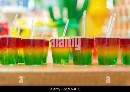 Glass with colorful striped jelly above wooden table. Rainbow jelly for kids party. Plastic spoon in jelly. Delicious Children's Party Dessert. Space Stock Photo