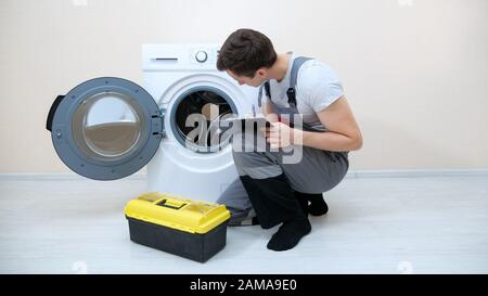 skilled repairman writes on clipboard checking broken washing machine on floor in room closeup Stock Photo
