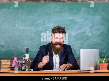 Teacher bearded man tell interesting story. Teacher interesting interlocutor as best friend. Teacher charismatic hipster sit table classroom chalkboard background. Telling educational stories. Stock Photo