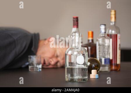 Drunken man sleeping with his head lying on a table and open mouth and alcohol bottles scattered around him. One bottle is flipped. Stock Photo