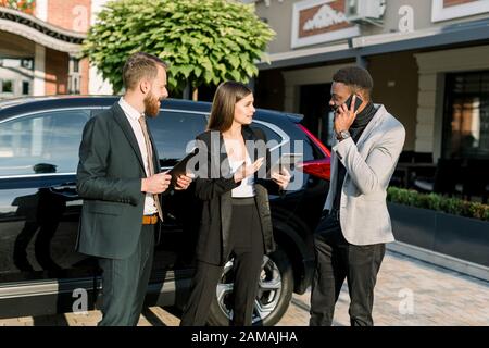 Three young multiethnical business people discuss something outdoors near black car. Caucasian women holds tablet, African man speaking on the phone Stock Photo