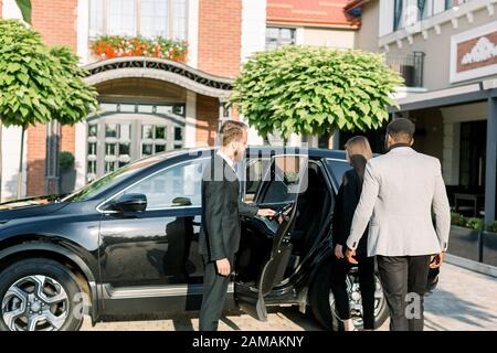 Chauffeur man opening car door for business couple, Caucasian woman and African man, side view. Business trip by car. Stock Photo