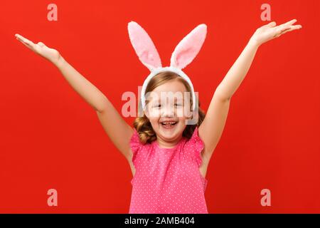 Happy easter. Cute little girl in bunny ears raised her hands up. Close-up baby on a colored red background. Stock Photo