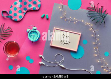 Frame with text 'Good night'. Healthy night sleep creative flat lay. Sleeping mask, blue mint alarm clock, earphones, earplugs and tea. Split pink and Stock Photo