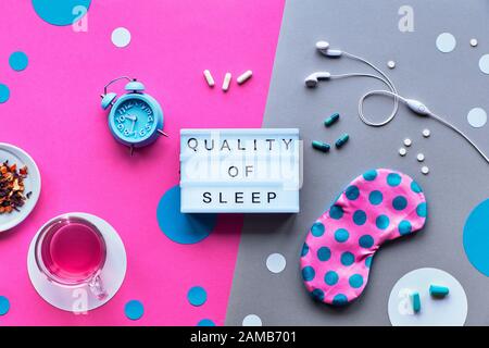 Text 'Quality of sleep' on trendy lightbox. Pink sleeping mask with polka dots, alarm clock, earphones and earplugs. Pills, capsules and calming tea. Stock Photo