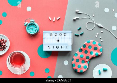 Text 'Good night' on trendy lightbox. Pink sleeping mask with polka dots, alarm clock, earphones and earplugs. Pills, capsules and calming tea. Flat l Stock Photo