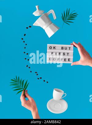 Trendy levitation. Flying line of coffee beans between ceramic coffee maker and espresso cup. Palm leaf and lightboard with text "OK but coffee first" Stock Photo