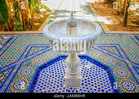 White stone fountain in the middle of green garden of Islamic palace. It's in Morocco. The fountain stands on blue-white tiles with typical Arab and Stock Photo