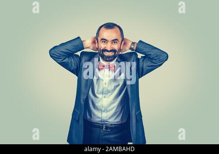 Happy, large smiling businessman keeping hands behind his head looking surprised at you camera. Human gestures. Happy man with dark suit, blue shirt, Stock Photo