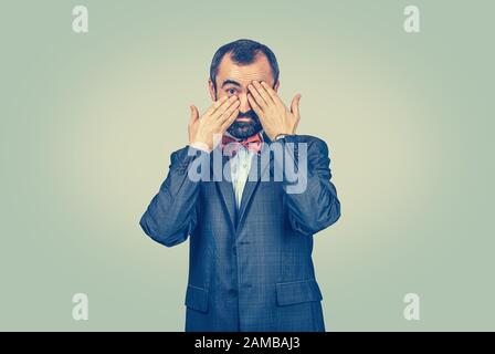 Handsome man with smile covering one eye with hands as he looks sneaky at the camera. Isolated, half length body, light green yellow background. Human Stock Photo