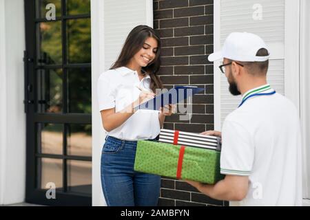 Side view of young bearded deliveryboy in white cap giving two gift boxes to woman. Pretty smiling brunette female in casual outfit receiving order and signing paper near house. Delivery concept. Stock Photo