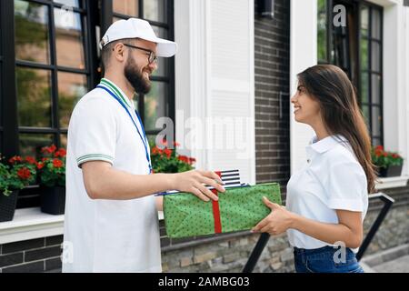 Side view of young bearded deliveryboy in white cap giving two gift boxes to woman. Pretty smiling brunette female in casual outfit receiving order and folder to sign near house. Delivery concept. Stock Photo