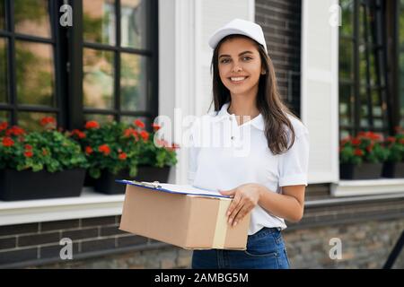 Front view of pretty happy joyful young female courier in white cap and casual outfit carrying box and folder. Brunette woman standing outdoors, holding parcel and smiling. Delivery concept. Stock Photo