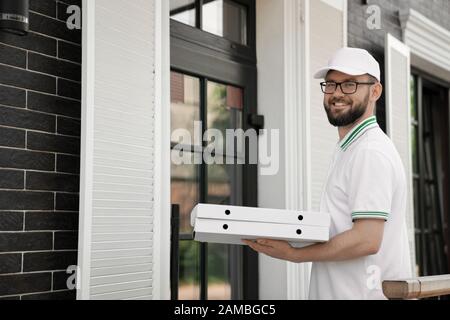 Side view of happy joyful young male courier in white polo and cap carrying pizza outdoors. Bearded man in glasses standing near door, holding two pizza boxes and smiling. Food delivery concept. Stock Photo
