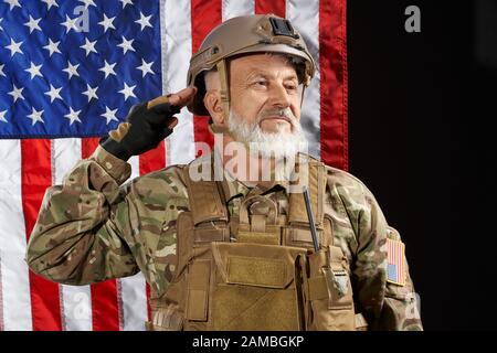 Front view of old military officer saluting and looking away. Portrait of bearded american veteran in uniform and helmet posing on black background with flag. Concept of military, army. Stock Photo