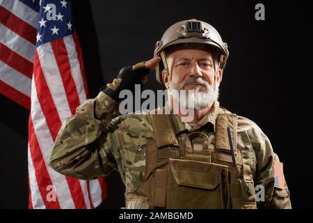 Front view of old military officer saluting and looking at camera. Portrait of bearded american veteran in uniform and helmet posing on black background with flag. Concept of military, army. Stock Photo