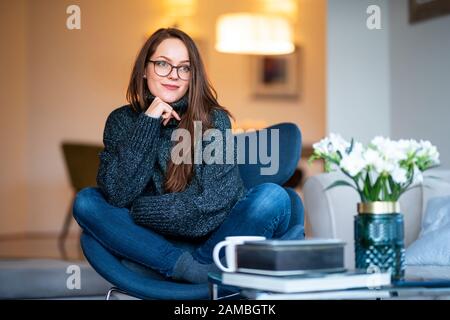 Portrait shot of smiling young woman daydreaming while relaxing on chair at home. Stock Photo