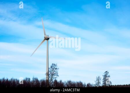 Three bladed wind turbine generator and pylon situated above Stracathro, Angus, Scotland, UK surrounded by trees, blades blurred revealing motion. Stock Photo