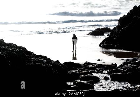 Silhouetted figure on wet sand on beach at Kynance Cove, Cornwall, UK, in black and white with high contrast