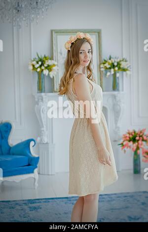 beautiful young girl in light dress and wreath of roses on head on International Women's Day is waiting for gift. Girl in beautiful white room against Stock Photo