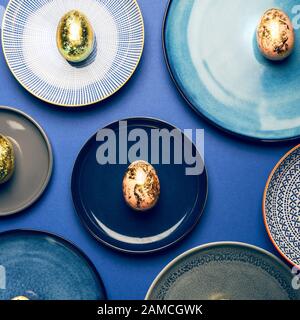 Different Ceramic Plates with Golden Speckled Easter Eggs of pastel colors on blue background. Happy Easter card concept, minimalistic design, top view Stock Photo