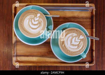 Top flat view of two cups of cappuccino with small heart and leave pattern of latte art in pale green ceramic cups on wooden tray and background. Stock Photo