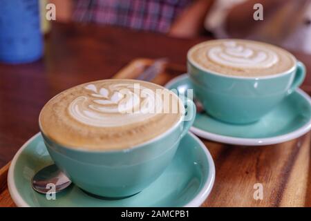 Two cups of cappuccino with small heart and leave pattern of latte art in pale green ceramic cups on wooden tray and blur background of cozy cafe. Stock Photo
