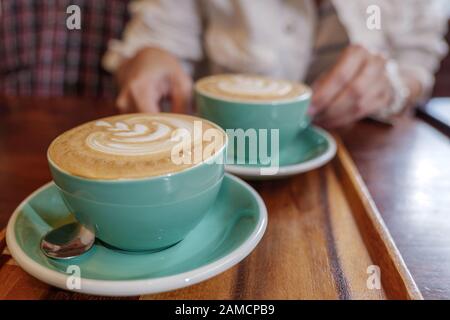 Two cups of cappuccino with small heart and leave pattern of latte art in pale green ceramic cups on wooden tray and blur background of cozy cafe. Stock Photo