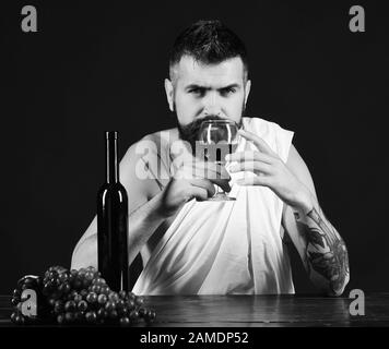 Winemaking and degustation concept. Sommelier smells drink. God Bacchus with serious face wearing white cloth sits by wine bottle and grapes. Man with beard holds glass of wine on brown background. Stock Photo
