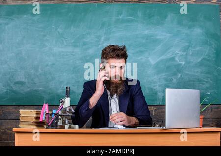 School principal or teacher calling parents to report about exam results. School teacher cares about communication with parents. Man with beard talk phone classroom background. In touch with school. Stock Photo