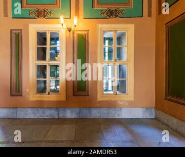 Corner orange wall with two narrow wooden window with closed green shutters, beautiful elegant rectangular green frames, and white tiled marble floor Stock Photo
