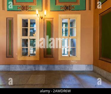 Corner orange wall with two narrow wooden window with closed green shutters, beautiful elegant rectangular green frames, and white tiled marble floor Stock Photo