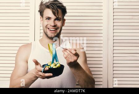 Fitness lifestyle and regime idea. Man with unshaven face holds bowl and fork with measuring tape. Weight management and sportive diet concept. Guy with happy face on background of beige jalousie. Stock Photo