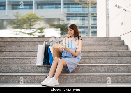 Beautiful Asian woman with shopping bags sitting on stairs while looking at smart phone and holding credit card. Stock Photo