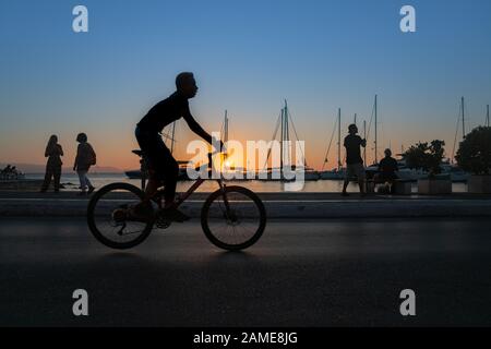 Naxos Greece - August 11 2019;  in silhouette a boy cycles past in foregropund while tourists look at view and locals fish in morning on Naxos town wa Stock Photo