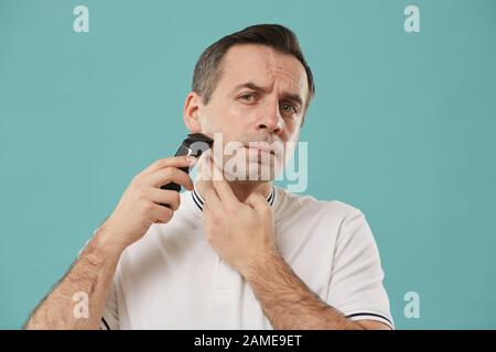 Head and shoulders portrait of handsome middle aged man shaving and looking at camera while enjoying morning routine standing against blue background Stock Photo