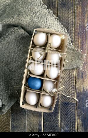 Eco-friendly wooden box with white eggs and one blue Easter egg on burlap on old wooden table. Zero waste packaging. Trendy color Classic Blue. Plasti Stock Photo