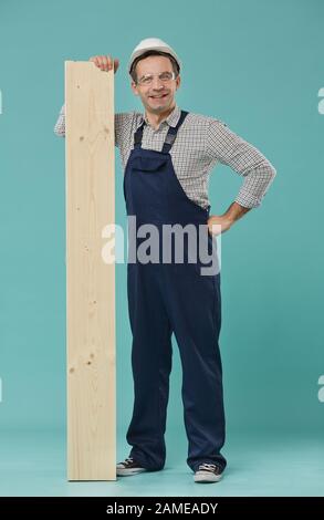 Full length portrait of professional worker or builder smiling at camera while leaning on wood standing against blue background Stock Photo