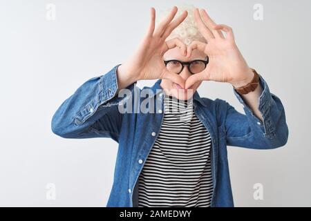 Young albino blond man wearing denim shirt and glasses over isolated white background Doing heart shape with hand and fingers smiling looking through Stock Photo