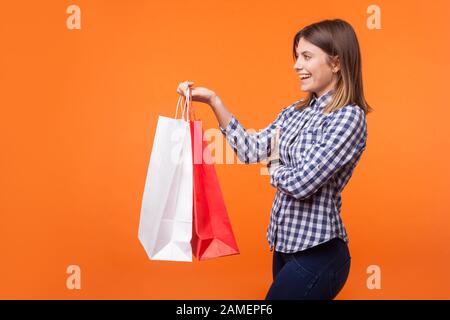 Side view portrait of friendly brunette woman with charming smile in casual checkered shirt standing giving shopping bags and smiling, offering goods. Stock Photo