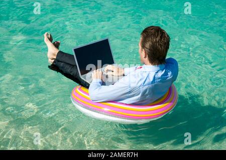 Relaxed businessman floating on a rubber ring while typing on his laptop Stock Photo