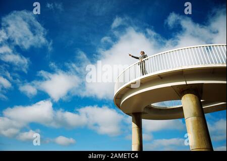 Businessman standing outdoors on curved walkway pointing into bright blue sky copy space Stock Photo