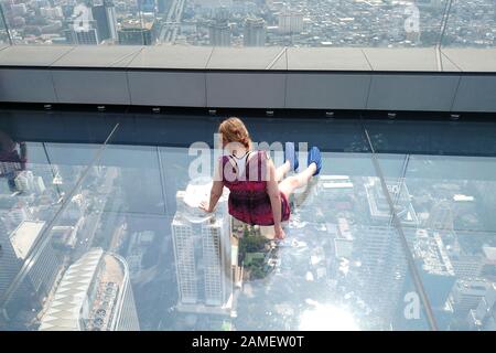 Bangkok, Thailand - December 21, 2019: Woman sitting on glass floor on rooftop of the King Power Mahanakhon building and looking down through glass. Stock Photo