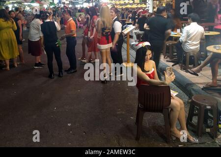 Busy street with lots of people walking and enjoying nightlife. Thai ladyboy with Christmas outfit sitting on chair and taking a selfie. Stock Photo