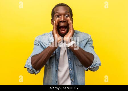 Attention! Portrait of scared man in denim shirt holding hands near wide open mouth and shouting announcement, looking with big eyes, shocked frighten Stock Photo